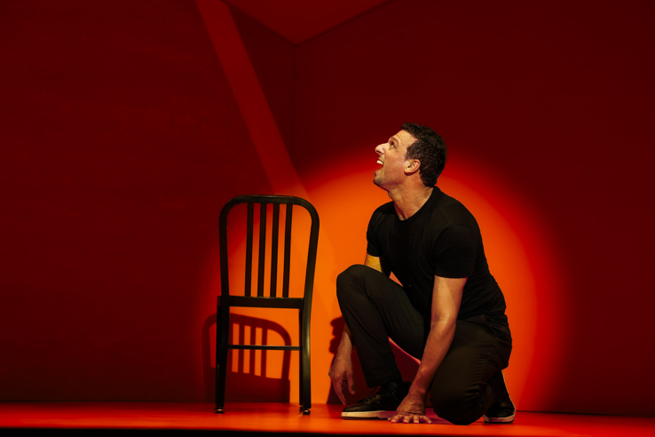 A man in al black crouching on the floor next to a chair and looking up