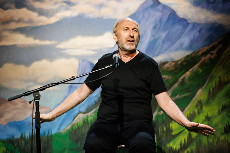 A photo of David Cale in a black shirt at a microphone talking with his arms outstretched