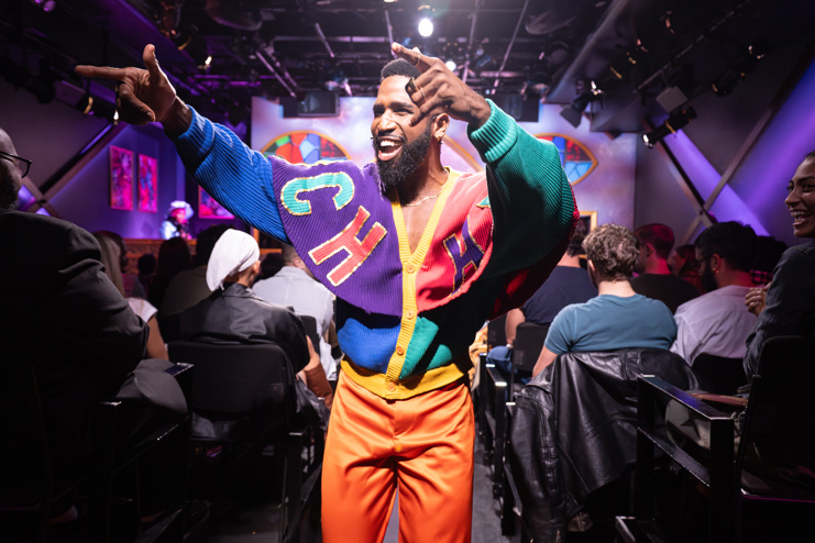 A man in a colorful sweater and orange pants walking through the audience smiling widely and pointing