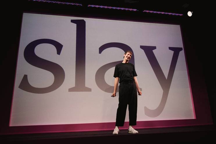 A man in all black standing onstage in front of a giant PowerPoint slide that says "slay"