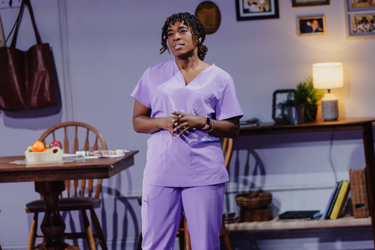 Kristolyn Lloyd onstage in purple scrubs looking pensive and standing in a modest living room