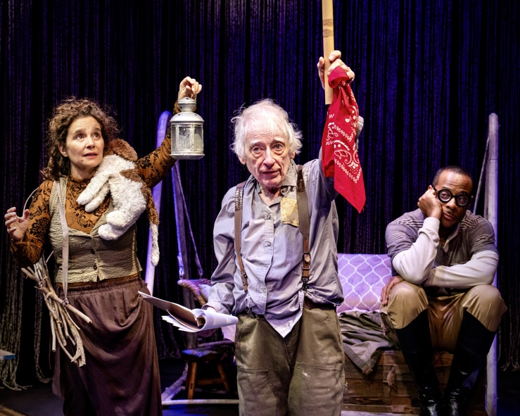 Austin Pendleton holding up a sword flanked by two other actors, one holding a lantern, the other looking bored