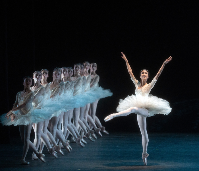 A ballerina in a white leotard and tutu on pointe with her arms aloft with a row of ballerinas in blue on the side