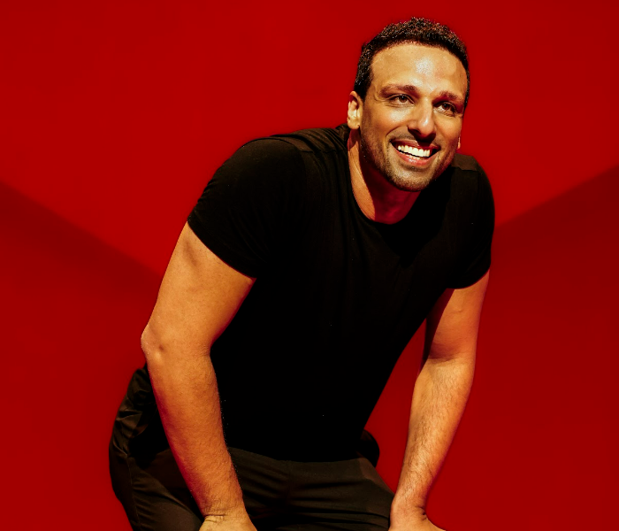 A man in a black T-shirt and pants bending slightly and smiling widely against an orange background