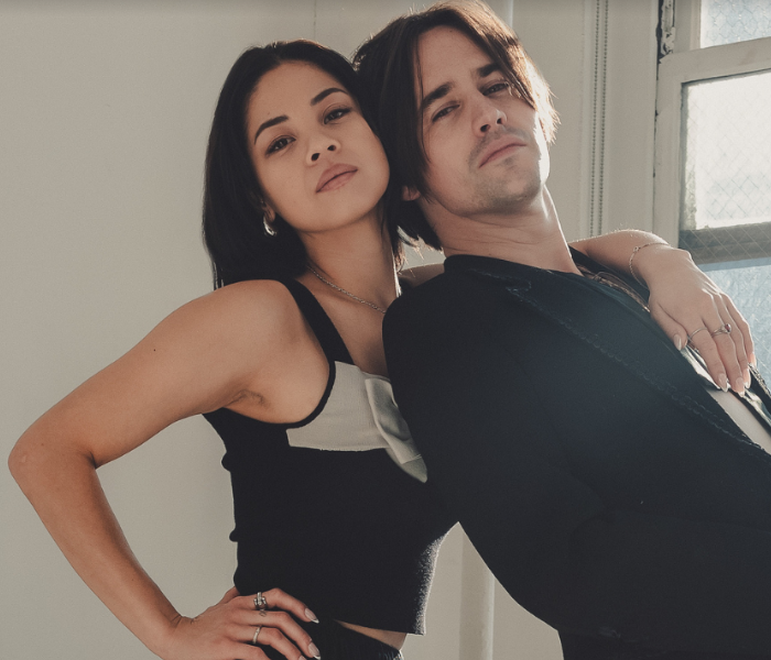 Eva Noblezada and Reeve Carney, with him leaning against her while she has her hand on her hip 