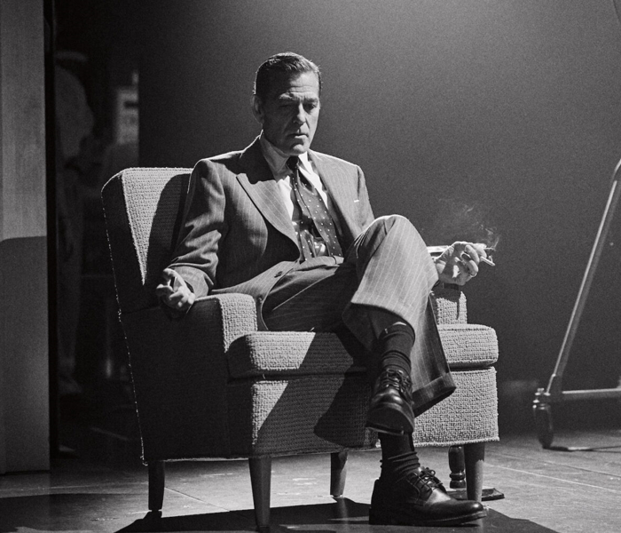 A black-and-white photo of George Clooney sitting in a chair smoking a cigarette looking pensive 