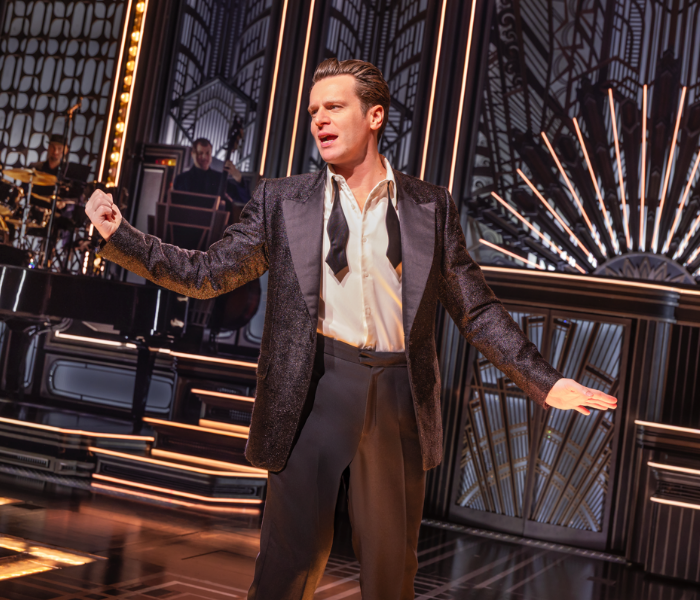 Jonathan Groff onstage in a suit with an open tie singing with his arms extended 