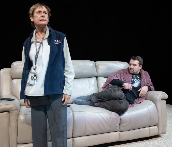 A middle-aged woman standing looking off concerns while a young man lounges on a couch behind her 