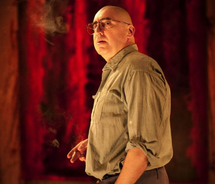 A bald Alfred Molina as artist Mark Rothko, looking serious in a khaki shirt against a red painting 